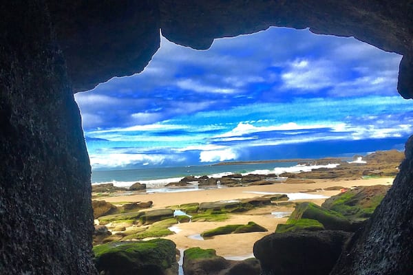 A rocky beach with a cave entrance, calm waters, and a cloudy sky.