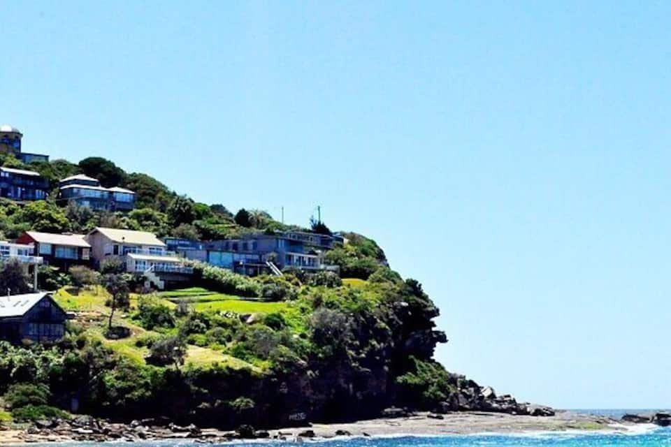 A beach with houses on a cliff, a person sitting on the sand, and clear blue water.