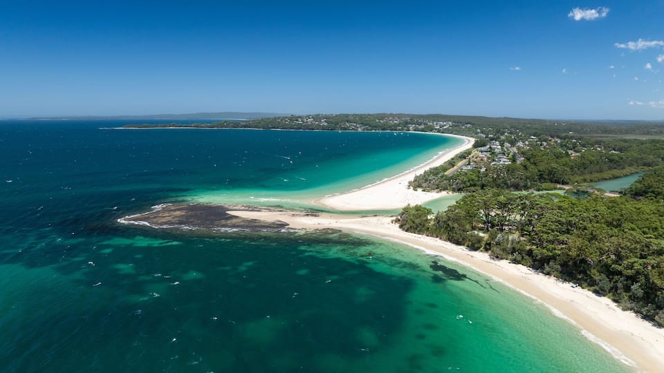 A coastal landscape with a sandy beach, clear turquoise waters, and a forested area.