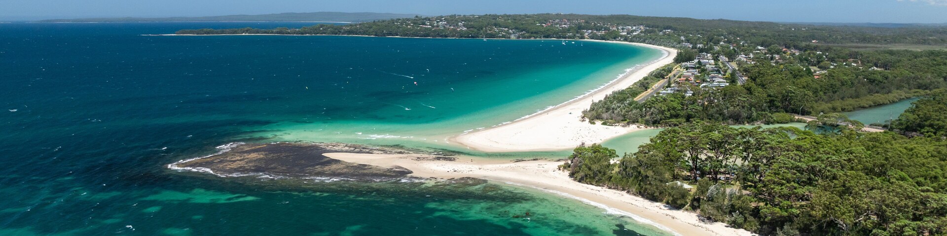 Paisagem costeira com uma praia arenosa, águas turquesa cristalinas e uma área arborizada.