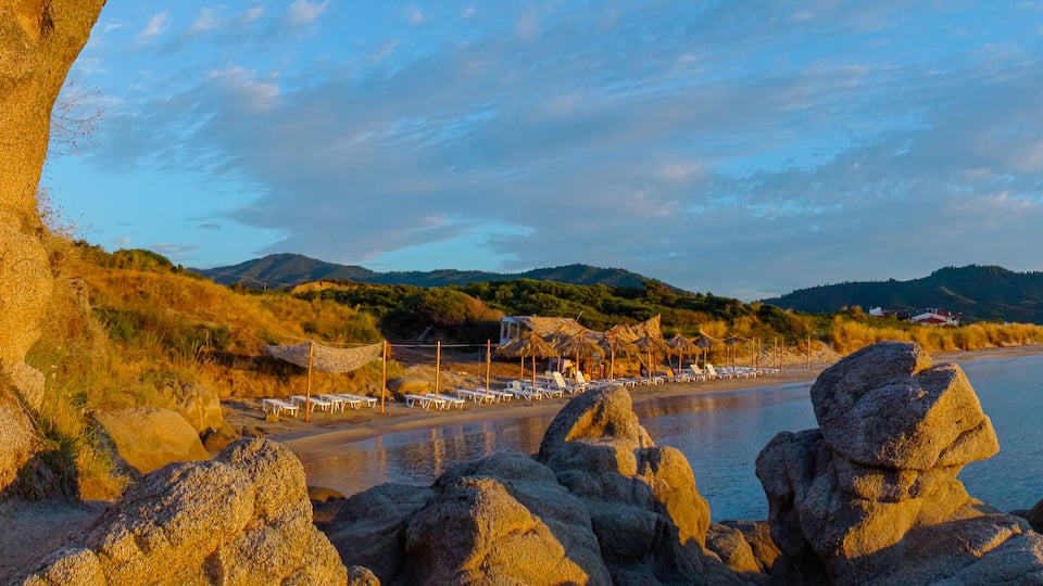 A coastal landscape with a rocky shoreline, a beach with sun loungers, and a backdrop of hills and a clear sky.