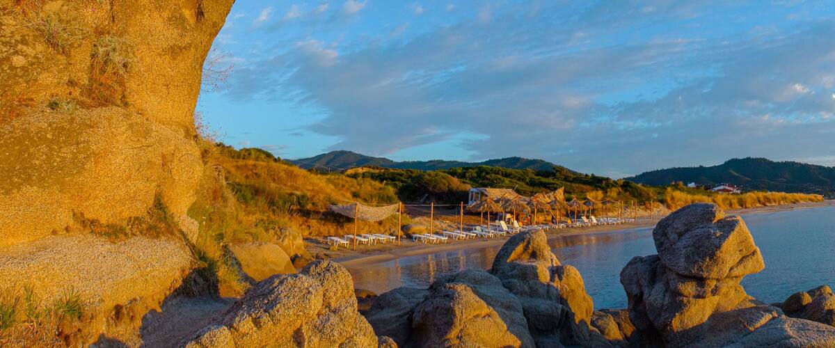 A coastal landscape with a rocky shoreline, a beach with sun loungers, and a backdrop of hills and a clear sky.