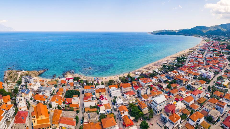 A coastal town with a beach, houses with orange roofs, and mountains in the background.