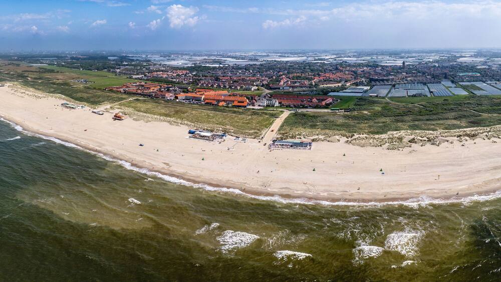 A panoramic view of Ter Heijde village shows an expansive sandy beach curving around a body of water, with buildings and farmland visible in the background.