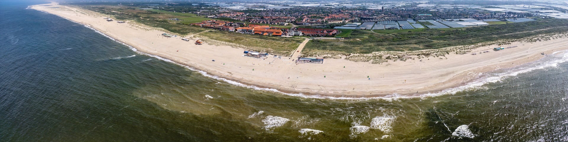 A panoramic view of Ter Heijde village shows an expansive sandy beach curving around a body of water, with buildings and farmland visible in the background.