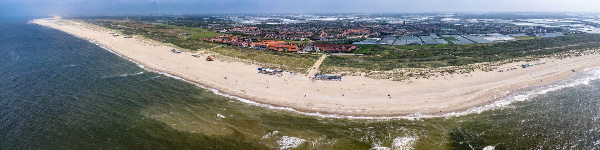 An aerial view of a coastal area with a sandy beach, a large building complex, and agricultural fields.