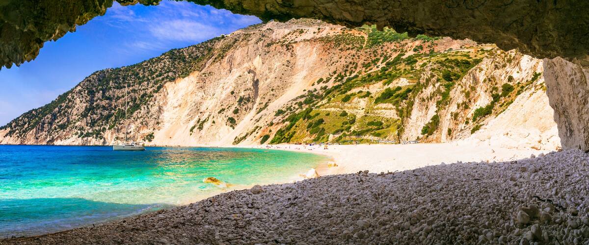 A coastal beach with clear turquoise waters, a rocky shoreline, and a mountainous backdrop.