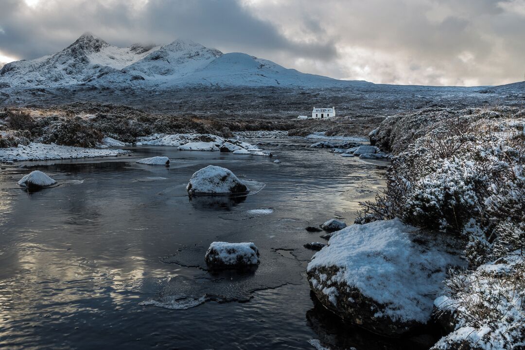 A snow-covered landscape with a river, rocks, and a white house in the distance.
