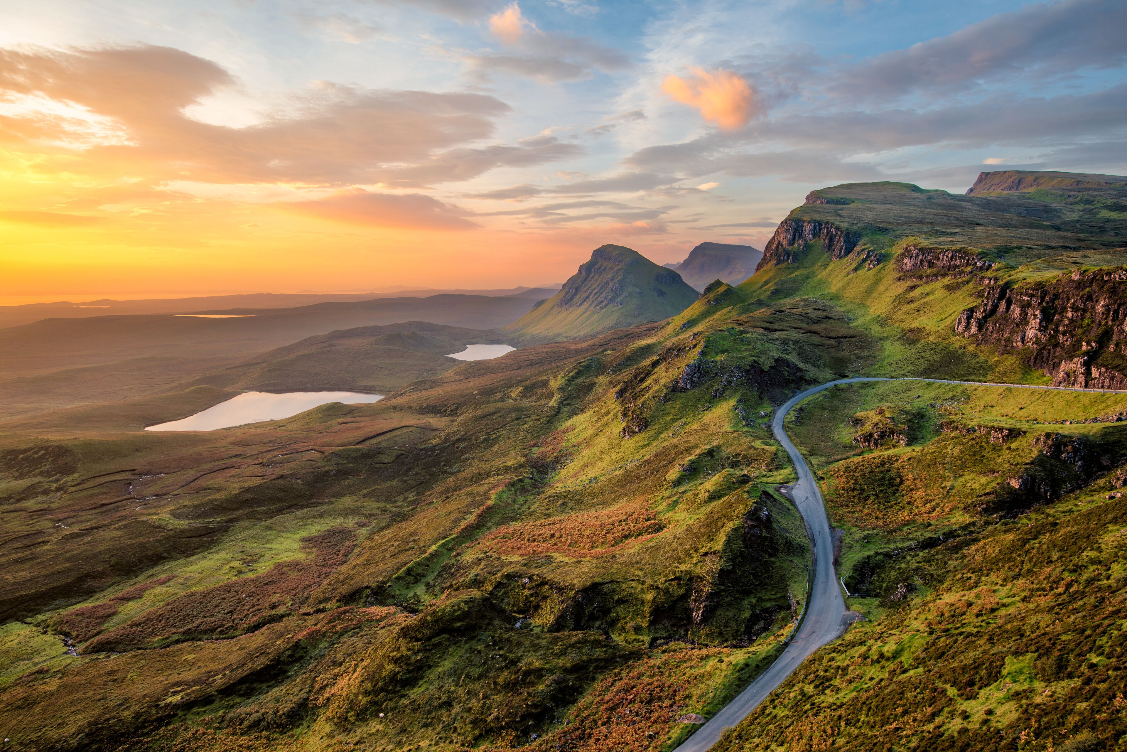 A winding road through a mountainous landscape with a lake and a sunset in the background.