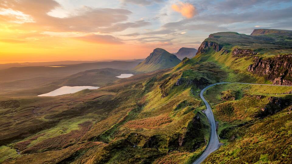 A winding road through a mountainous landscape with a lake and a sunset in the background.
