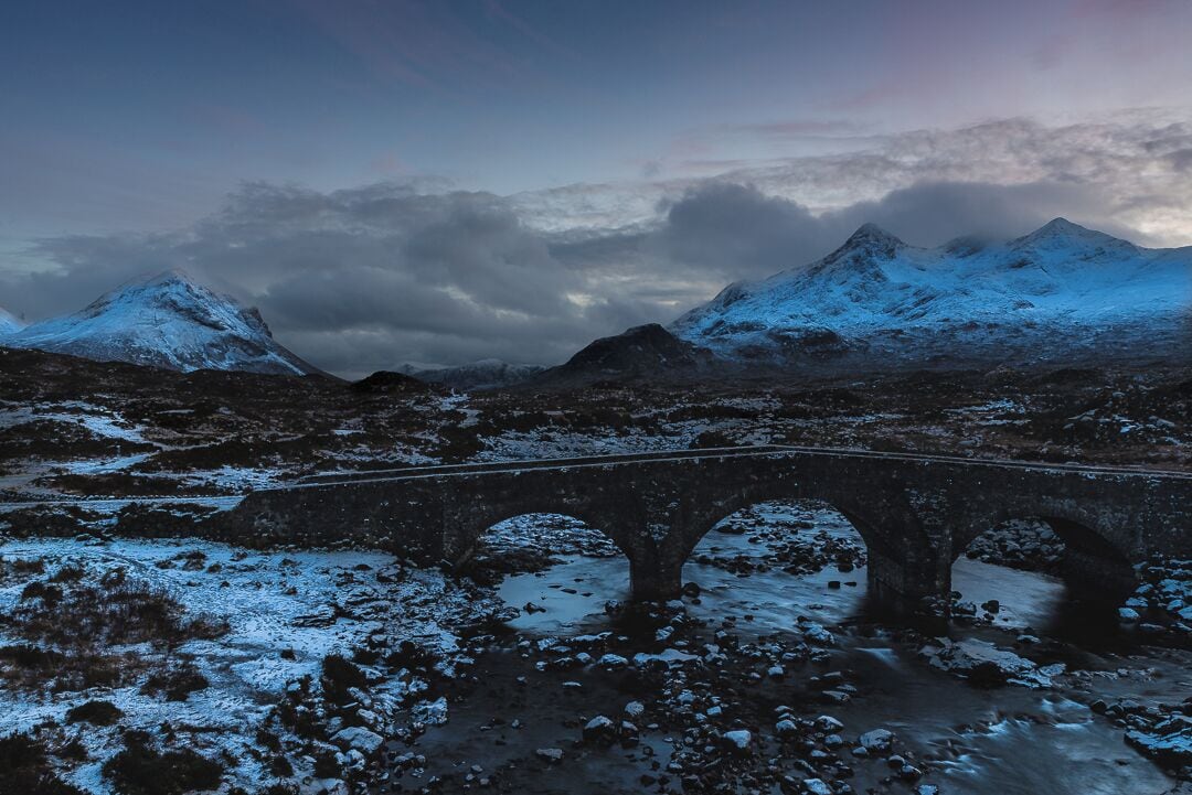 A snow-covered mountain range with a stone bridge in the foreground.