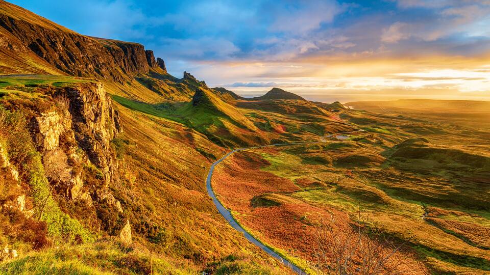 A winding mountain road through a valley with rocky cliffs and grassy terrain.