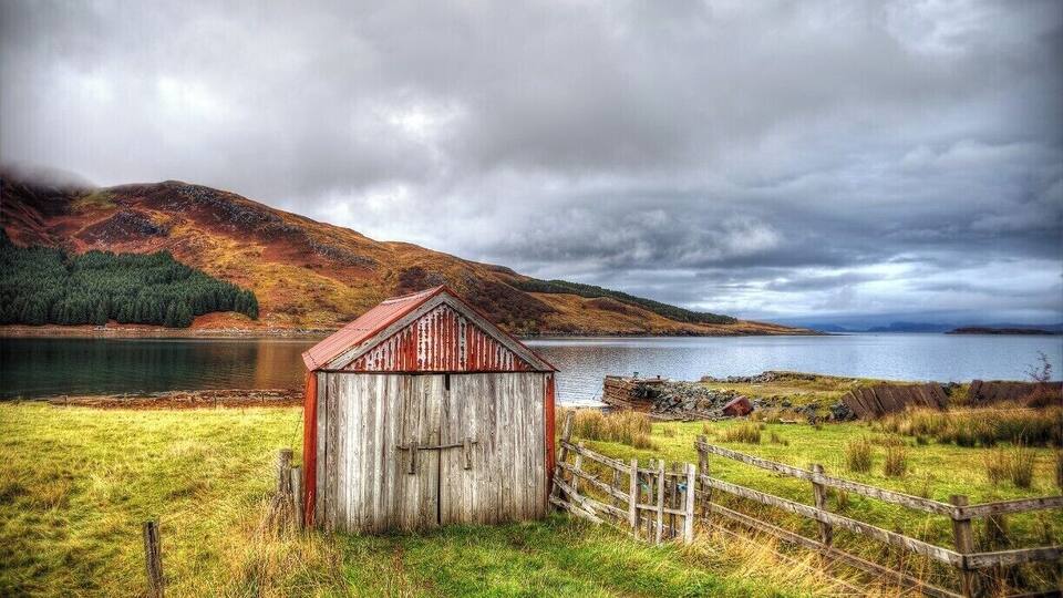 A rustic wooden shed by a lake with a wooden fence, surrounded by grassy fields and a forested hill.