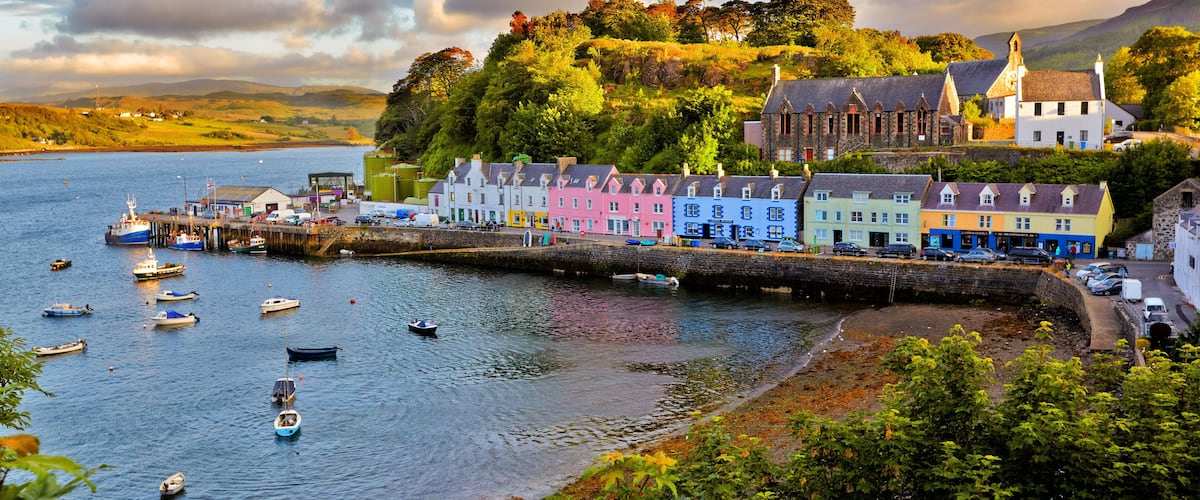 A coastal town with colorful houses, a pier, boats, and a hill with trees.