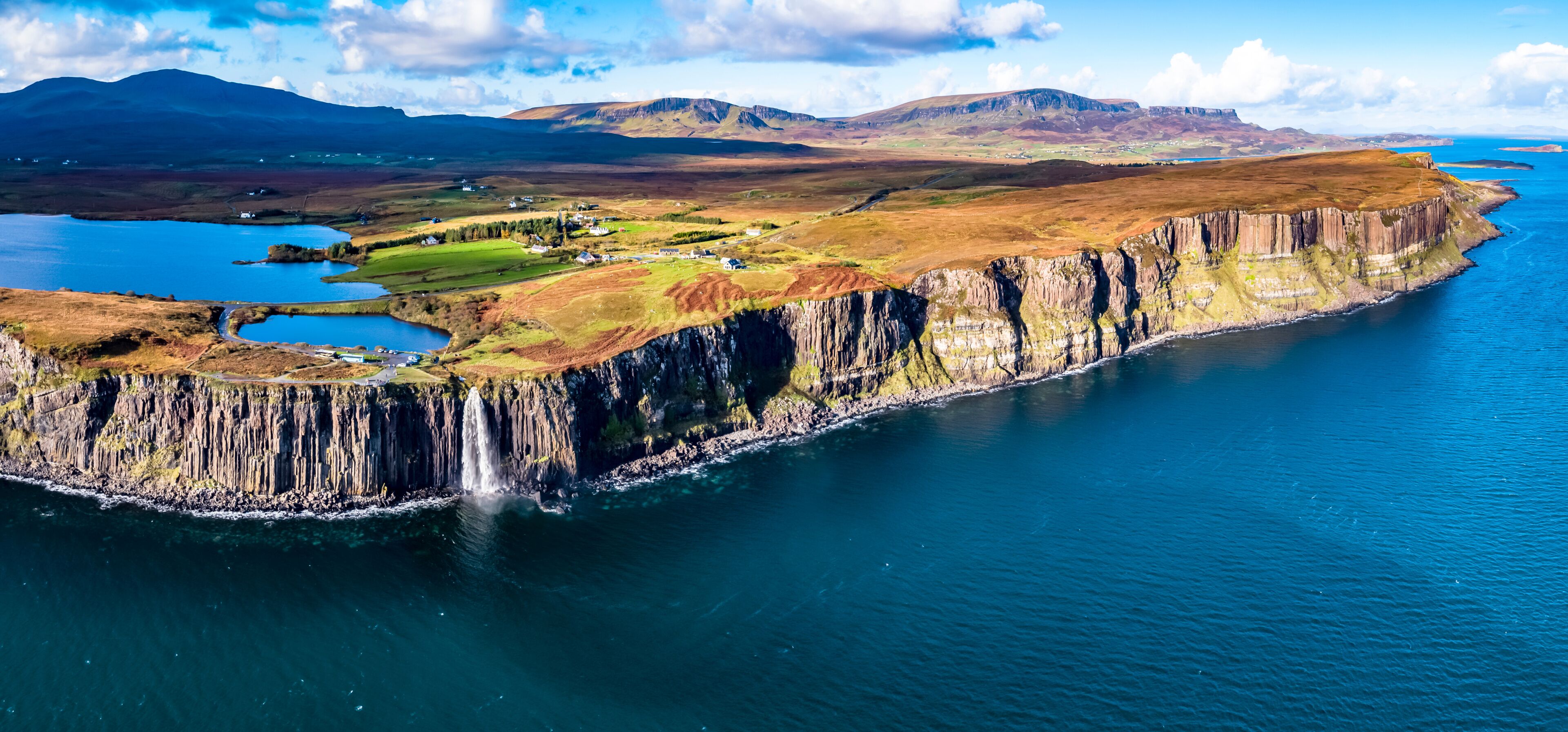 A coastal cliff with a waterfall, a small lake, and a distant mountain range.