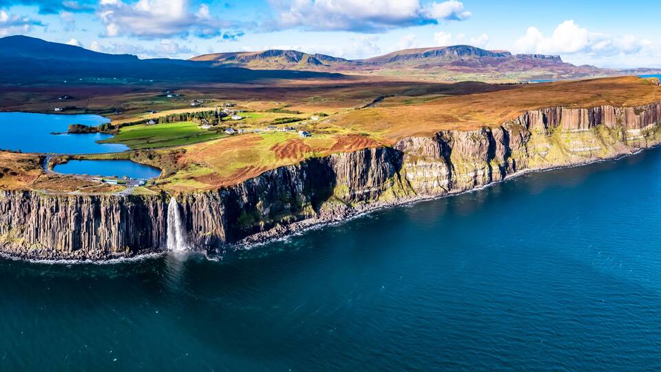 A coastal cliff with a waterfall, a small lake, and a distant mountain range.
