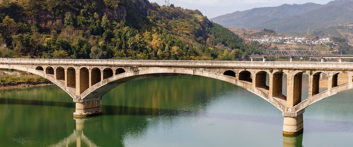 A stone bridge over a calm river with a forested hillside in the background.