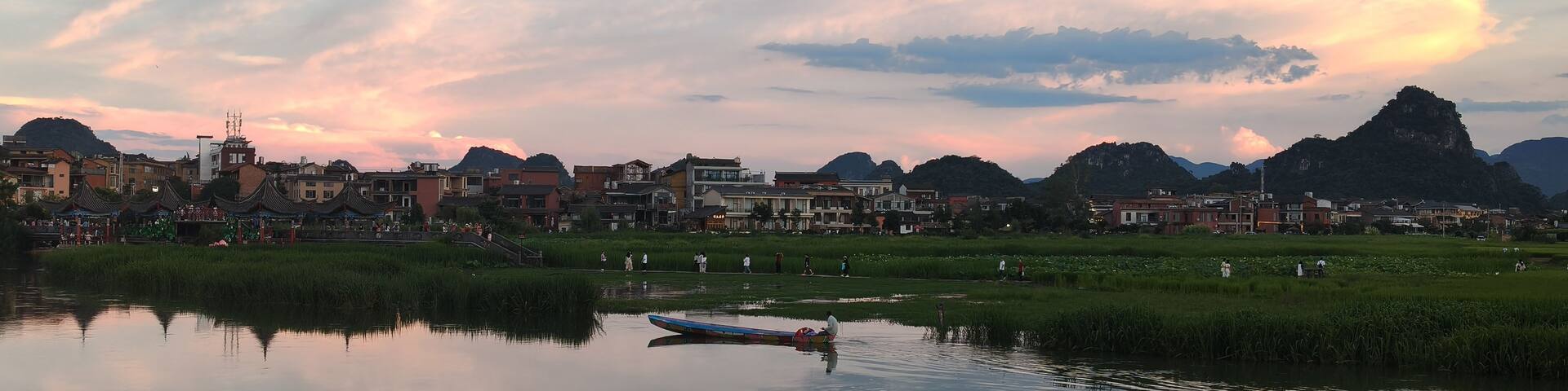 A calm river reflecting the sky, a traditional boat, and a town with buildings and mountains in the background.