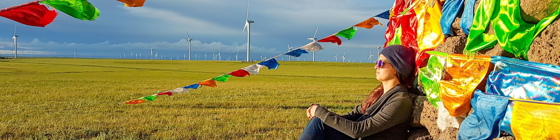 Una persona sentada sobre una roca, con coloridas banderas de oración y ruecas de oración en un campo de hierba, con aerogeneradores al fondo.