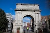 A stone archway with inscriptions, surrounded by buildings and a clear blue sky.