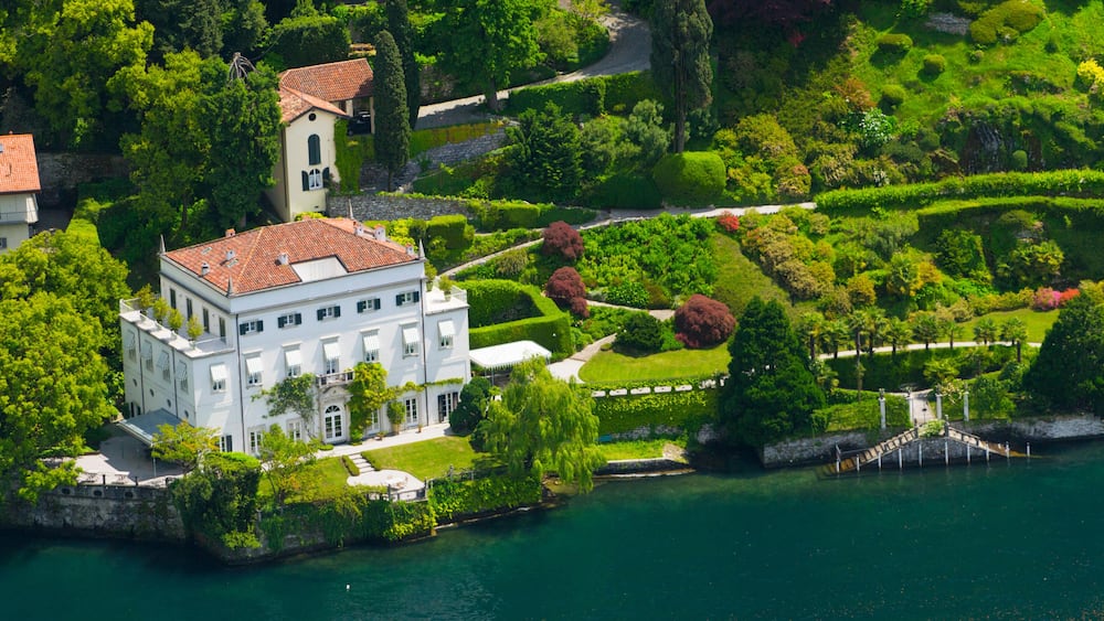 A white building with a red roof surrounded by lush greenery and a body of water.