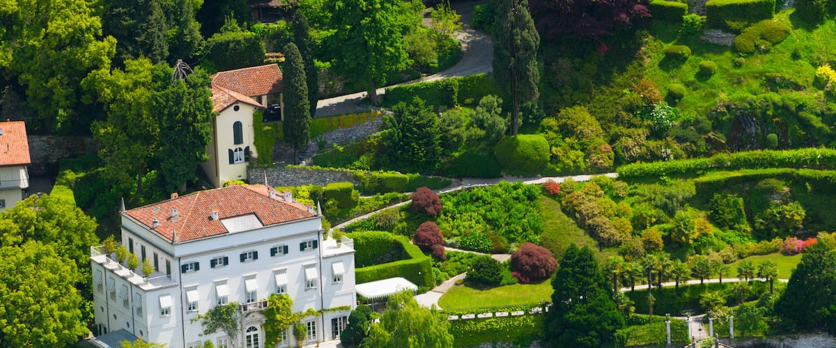 A white building with a red roof surrounded by lush greenery and a body of water.