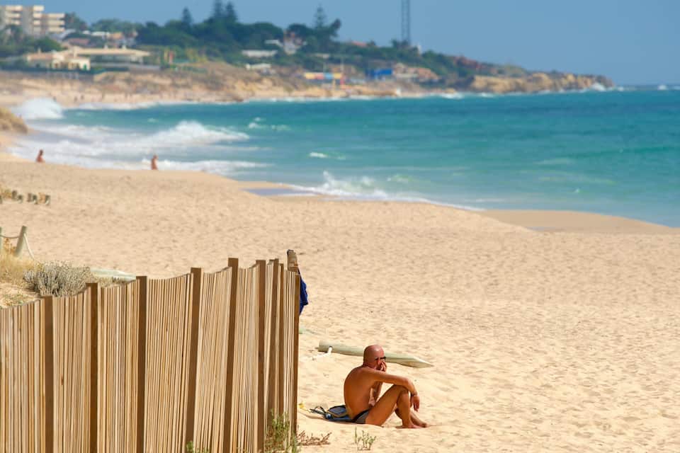 En sandstrand med ett trästaket, en person som sitter på sanden och havet i bakgrunden.