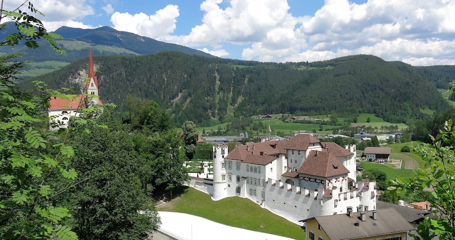 Un castello con tetto in tegole rosse, circondato da una vegetazione rigogliosa e montagne sullo sfondo.
