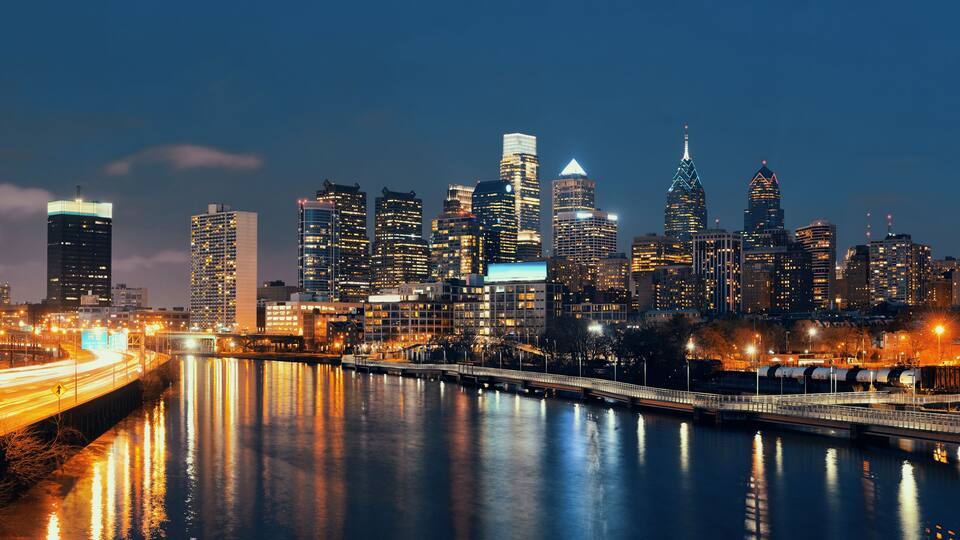 A nighttime city skyline with illuminated buildings, a river reflecting lights, and a bridge crossing the river.