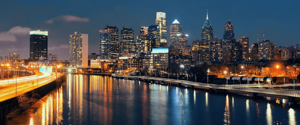 A nighttime city skyline with illuminated buildings, a river reflecting lights, and a bridge crossing the river.