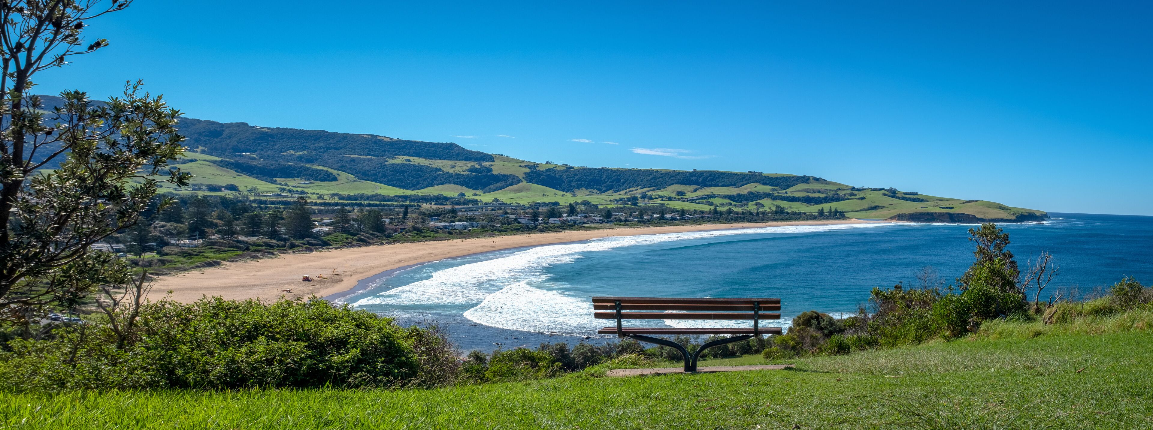 A coastal landscape with a bench overlooking the beach, green hills, and a clear blue sky.