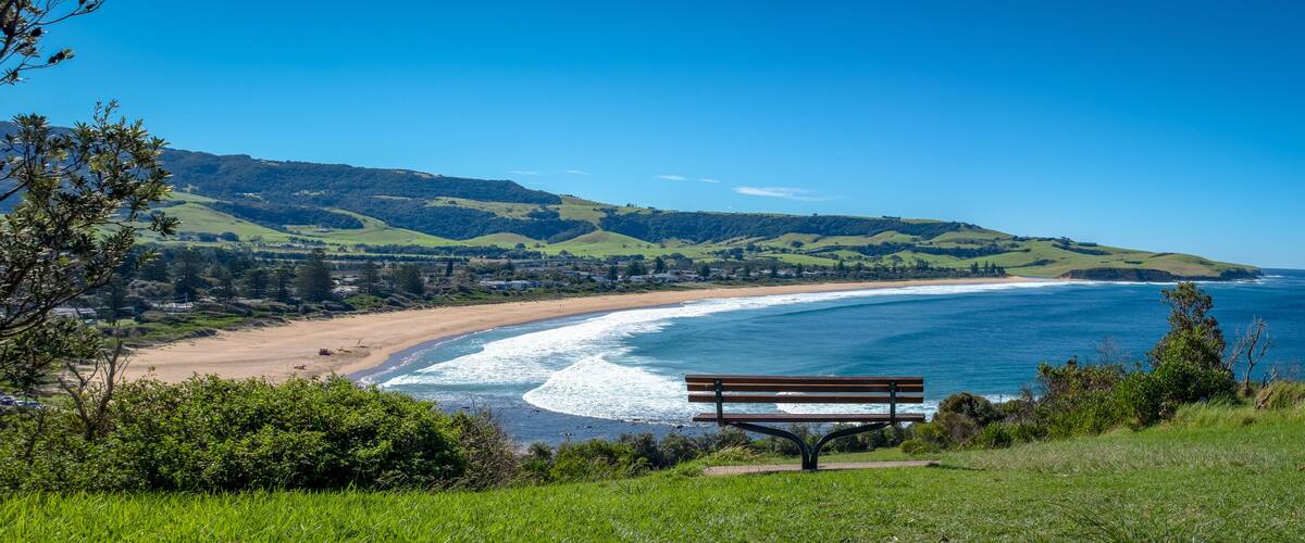 A coastal landscape with a bench overlooking the beach, green hills, and a clear blue sky.