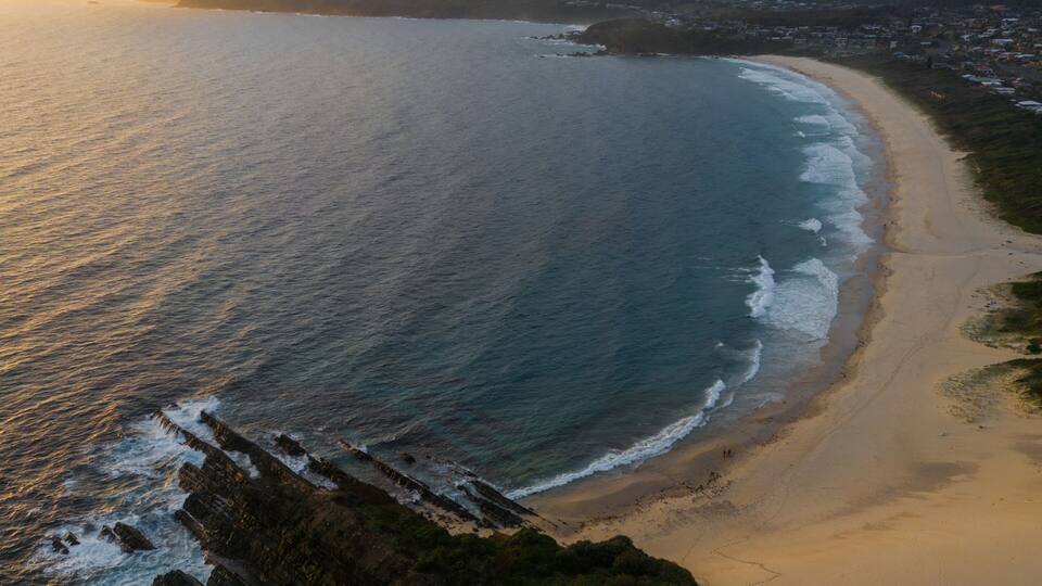 A coastal landscape with a sandy beach, rocky coastline, and a distant mountain range.