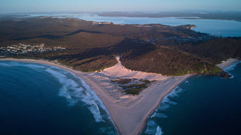 An aerial view of a coastal landscape with a sandy beach, a forested peninsula, and a bay with boats.