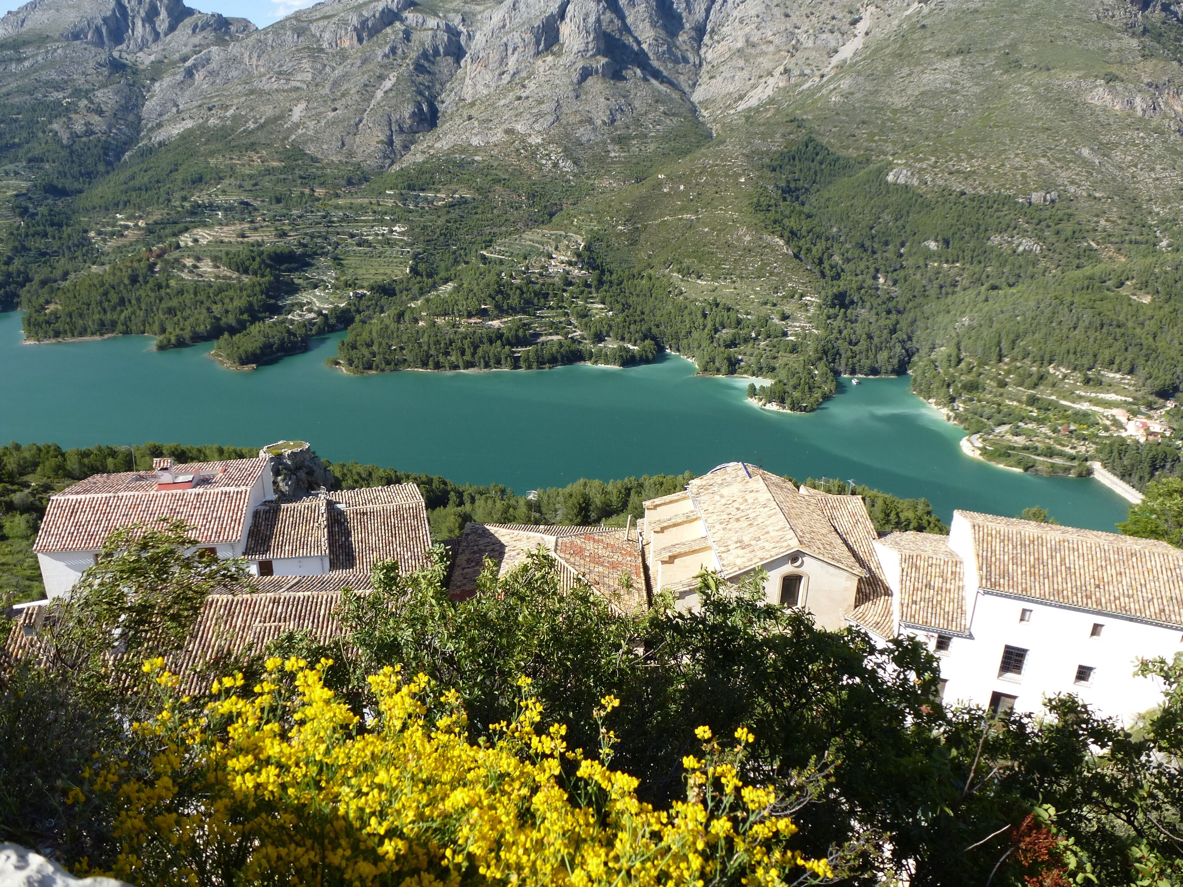 A mountainous landscape with a turquoise lake surrounded by greenery and buildings with terracotta roofs.