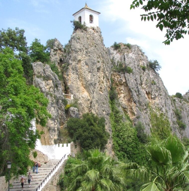 A white bell tower on a rocky cliff with a staircase leading up to it, surrounded by greenery.