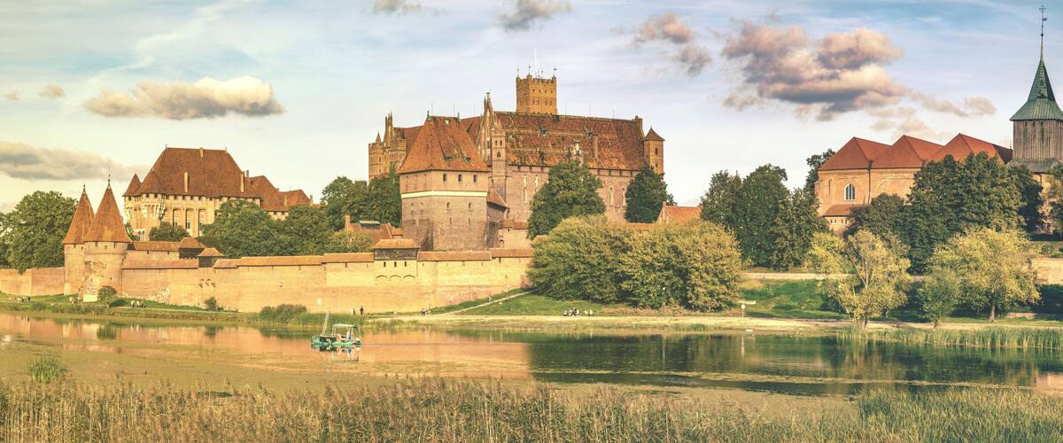 A historic castle with a moat, a church with a green spire, and a river with a boat.