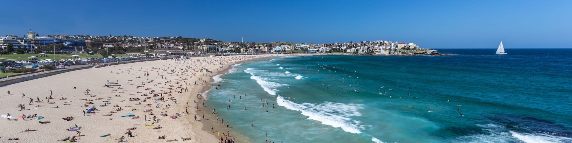 A beach with clear turquoise waters, a sailboat in the distance, and a cityscape in the background.