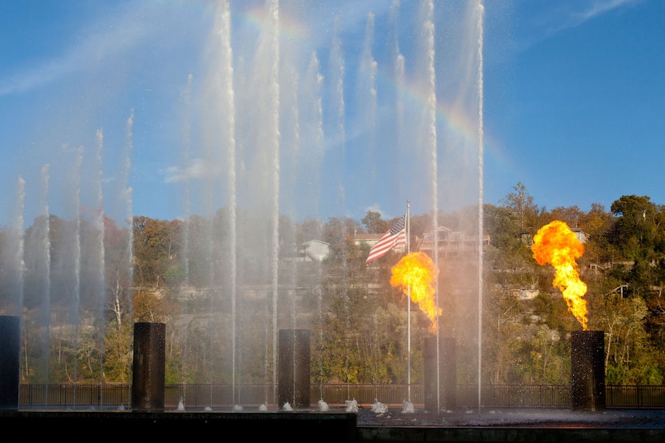 A fountain with water jets and a fire display, an American flag, and trees in the background.