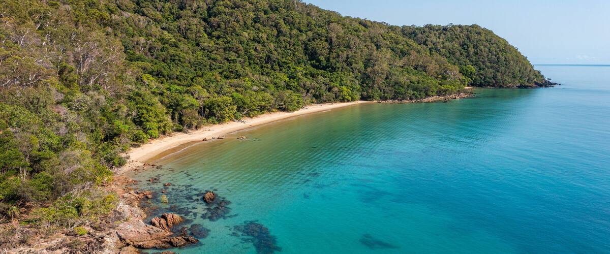 A coastal landscape with a sandy beach, clear turquoise waters, and a densely forested hill.
