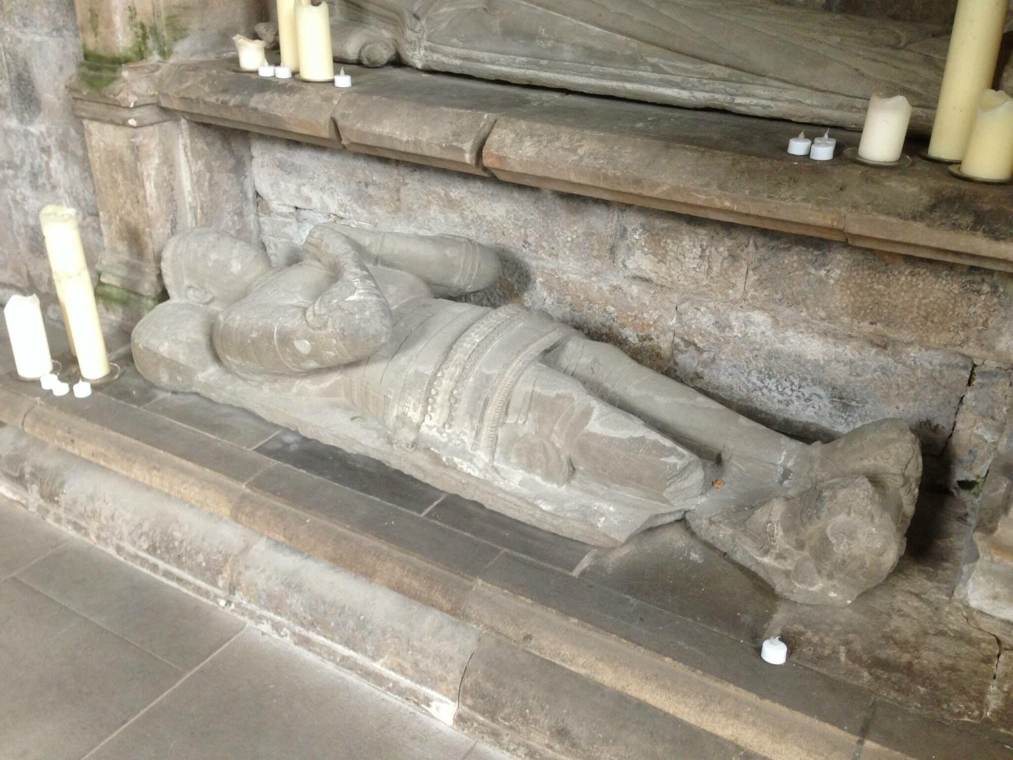 A stone sarcophagus with a reclining figure, candles, and a stone wall.