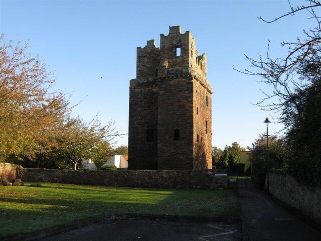 A historic stone tower with a narrow base and a wider top, surrounded by a grassy area and trees.