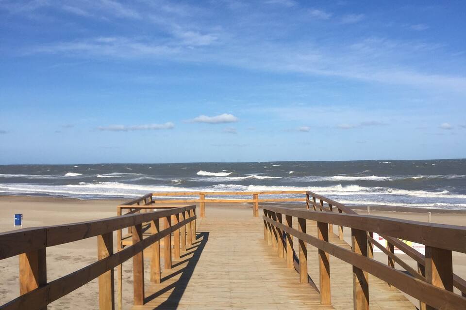 Un camí de fusta que condueix a una platja amb onades que trenquen a la riba sota un cel serè.
