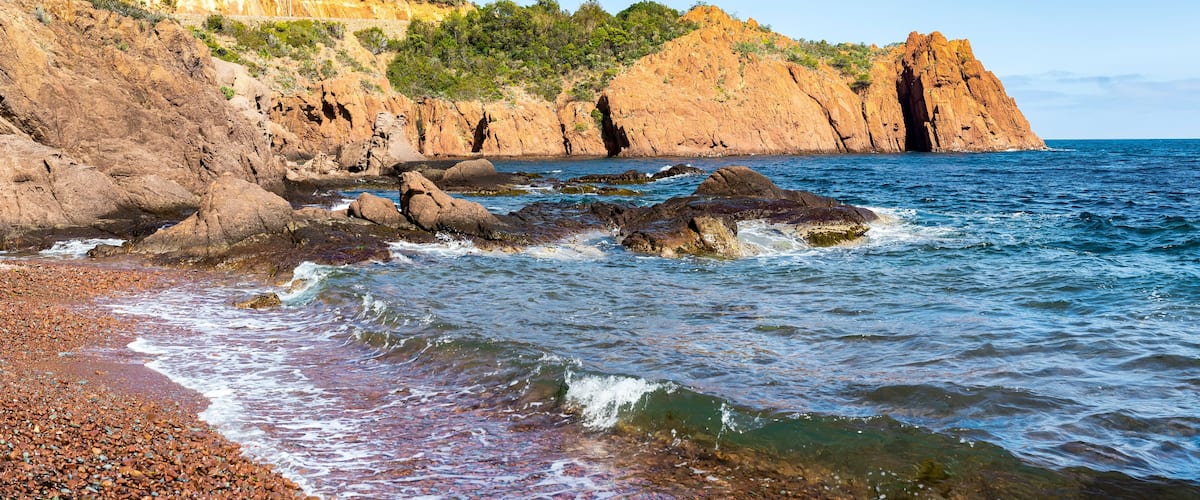A rocky coastline with clear blue waters, a pebbly beach, and a forested hill in the background.