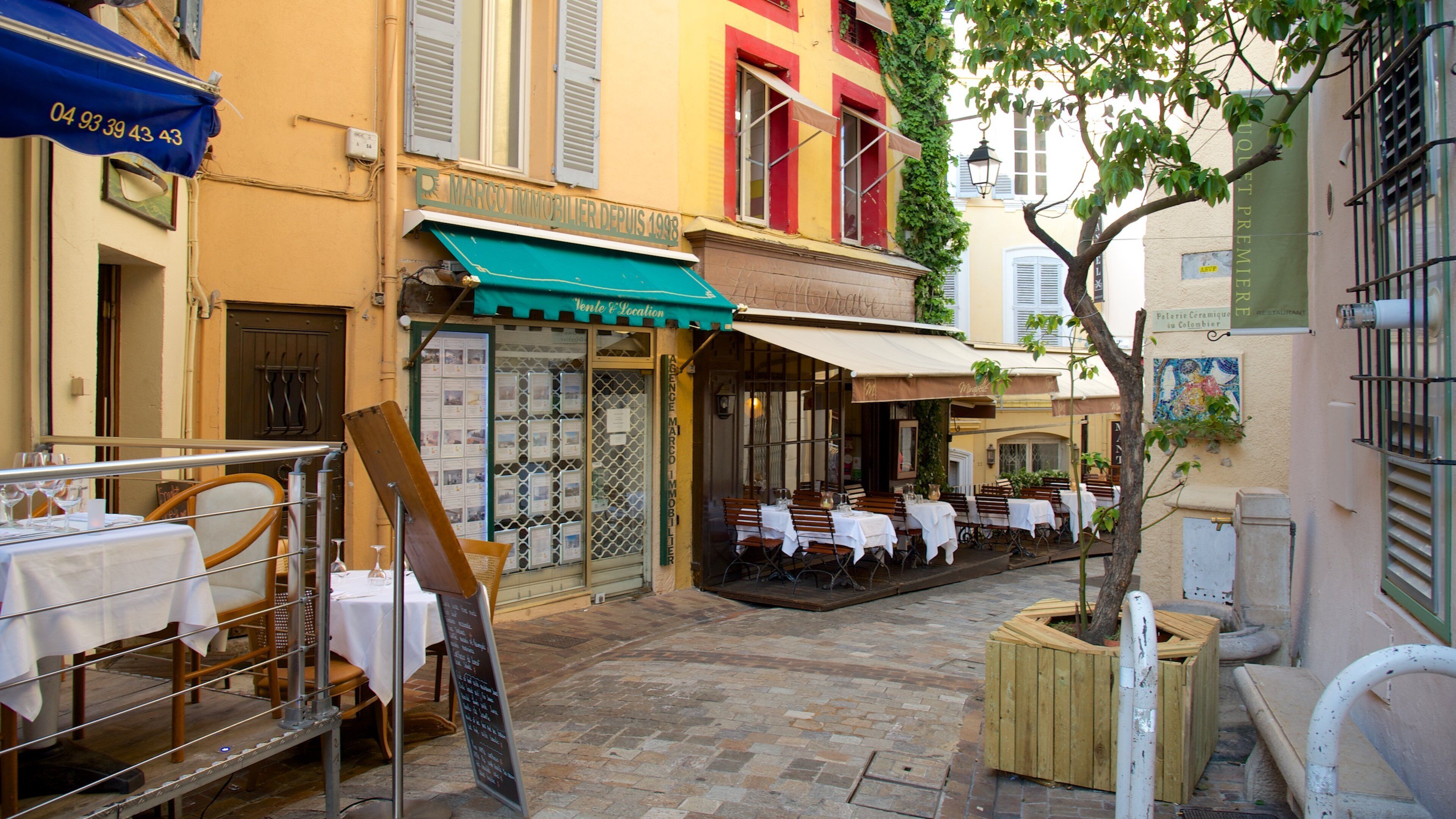 Una calle estrecha con mesas al aire libre en un restaurante, un toldo azul y un anuncio de una agencia inmobiliaria.
