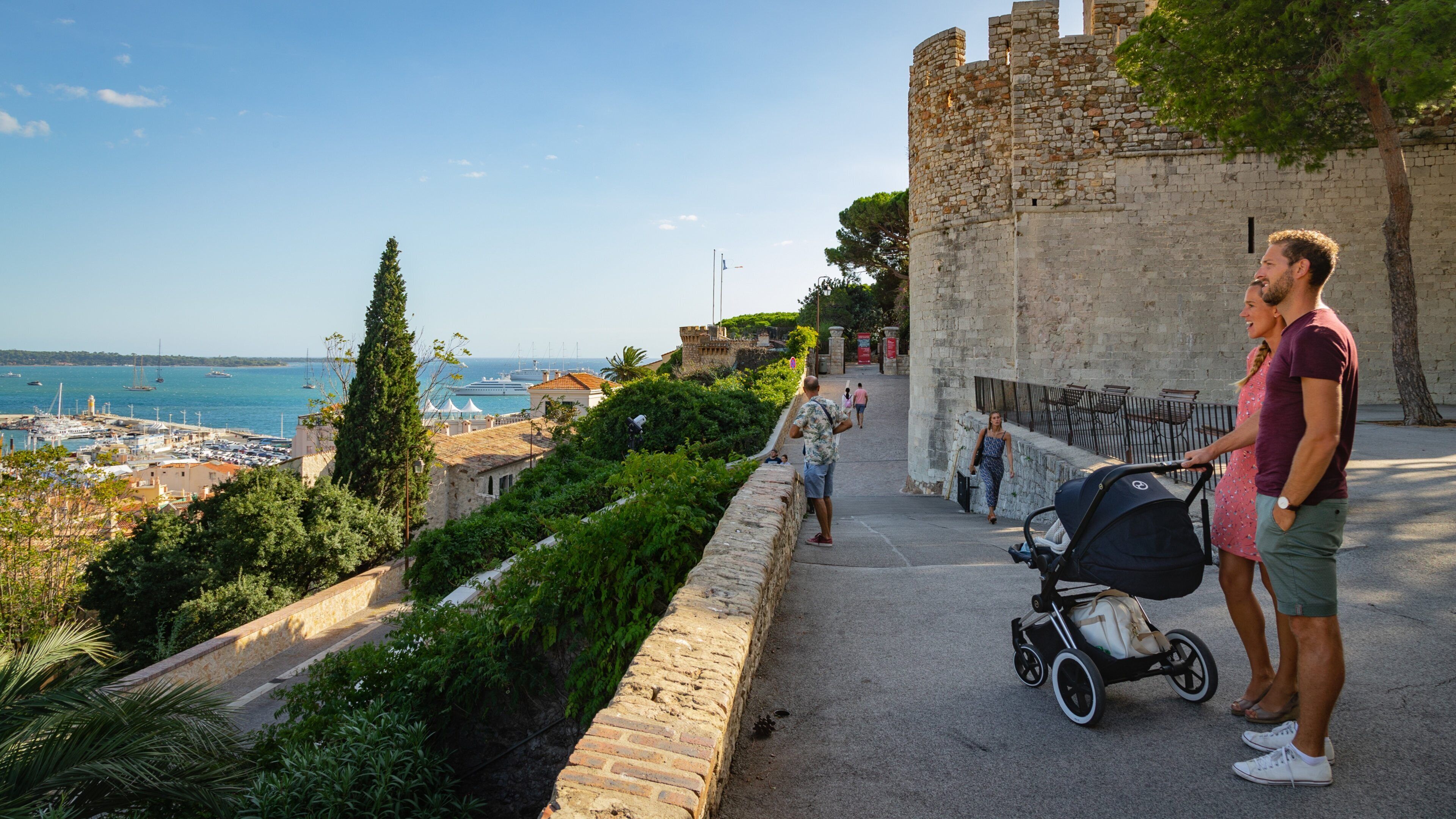 Uma vila costeira com uma muralha de pedra histórica, um porto com barcos e um céu azul límpido.