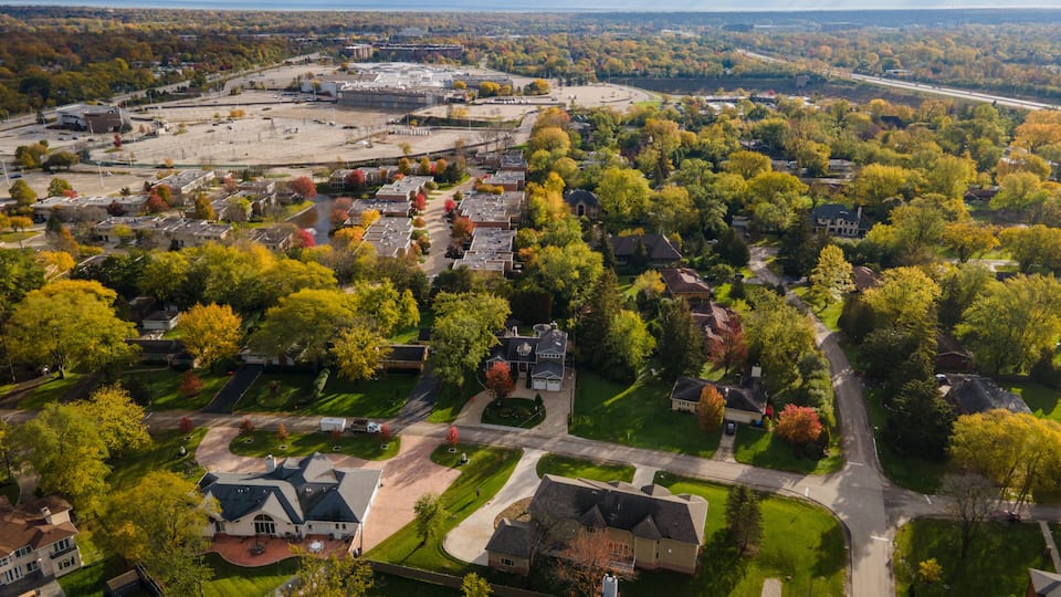 An aerial view of a suburban area with a large parking lot, residential houses, and a mix of deciduous trees.