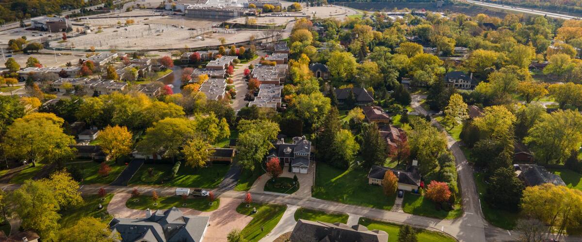 An aerial view of a suburban area with a large parking lot, residential houses, and a mix of deciduous trees.
