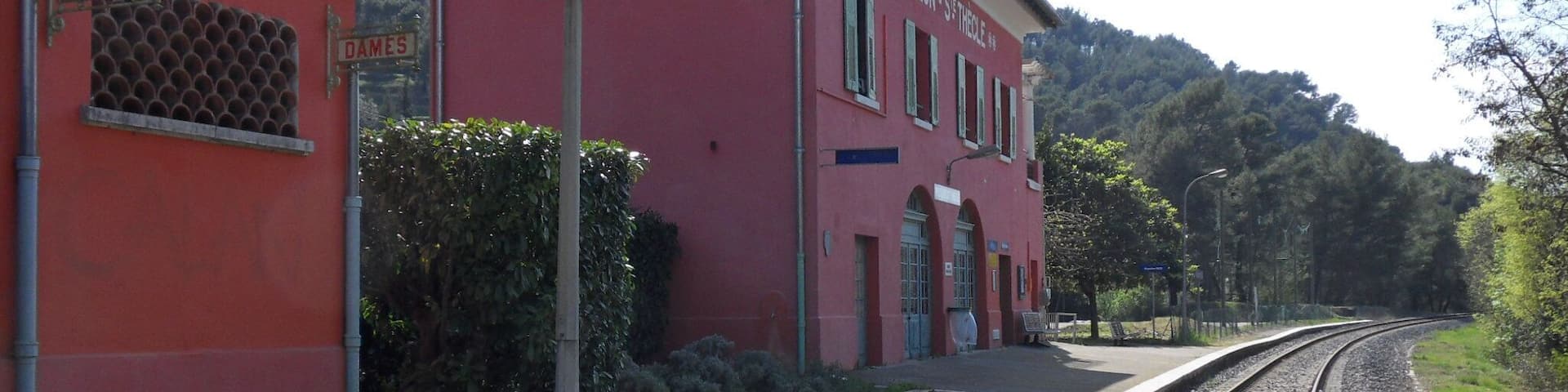 A red building with 'PEILLON ST THÈCLE' signage, a railway track, and a forested hill in the background.