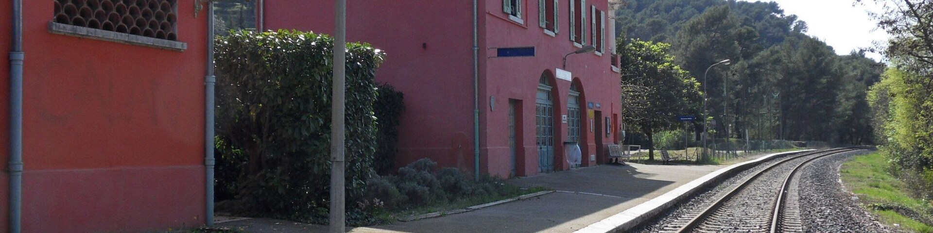 Un edificio rojo con señalización que dice 'PEILLON ST THÈCLE', una vía de tren y una colina boscosa al fondo.
